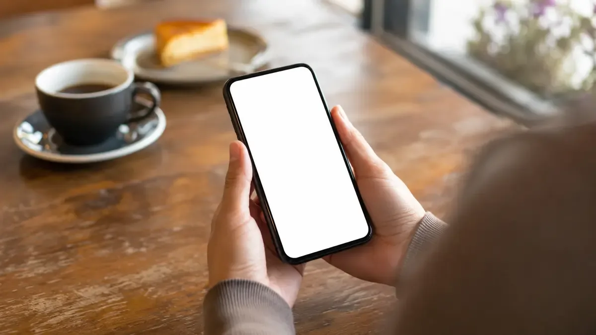 Manos sosteniendo un telefono celular de pantalla en blanco sobre una mesa de cafe